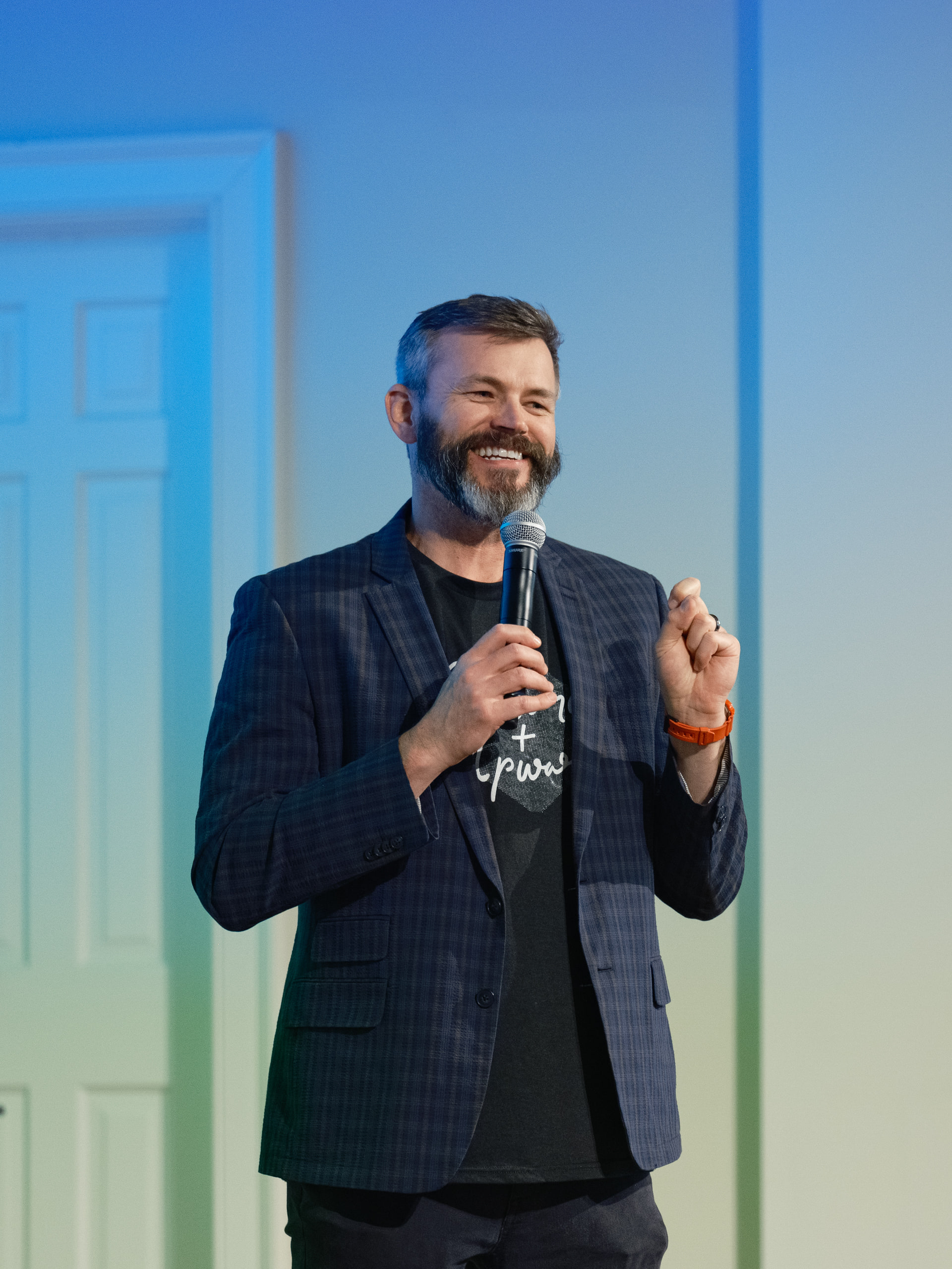 " A bearded speaker mid-sentence with a mic, navy plaid blazer and an orange watch, against a blue-to-green projected wall. Contrast, framing, color and subject clarity all four at once, which is exactly the brief.