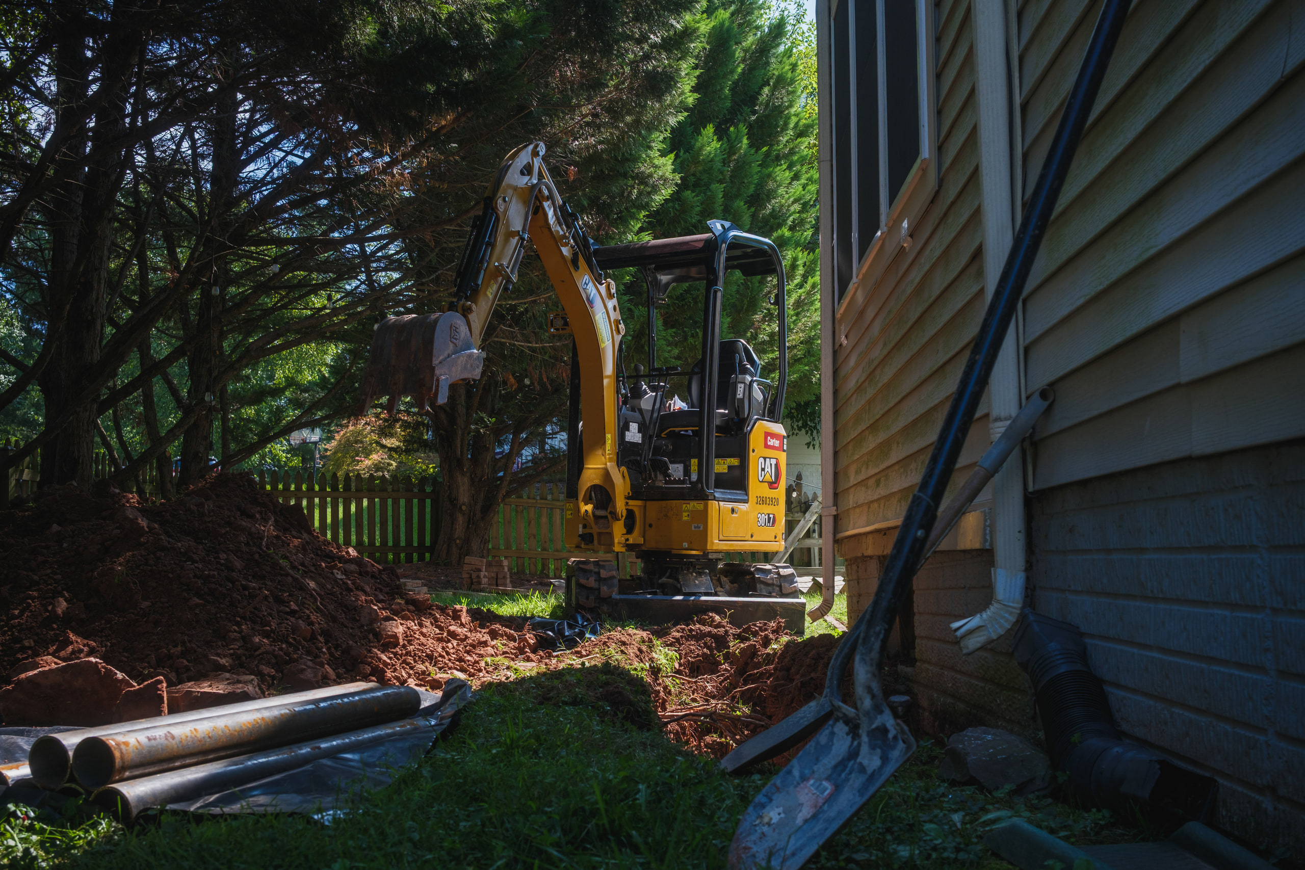 Ashburn location i just did... go for artsy... use the excavator instead." A CAT mini-excavator working the trench beside the house, dappled light. Landscape.